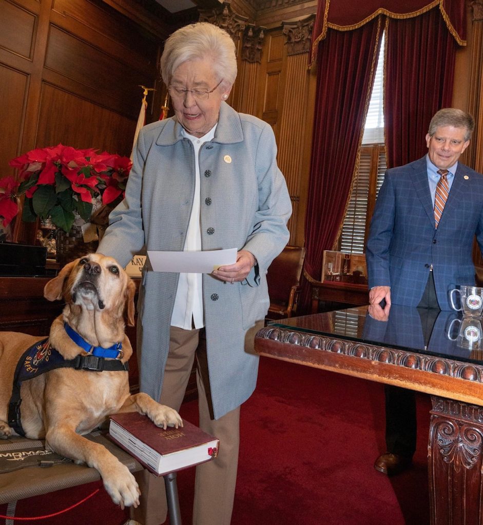 Millie, a yellow Labrador Retriever, looks up at her owner with her left paw on the Bible as Alabama Governor Kay Ivey pats her head and reads off a sheet of paper.