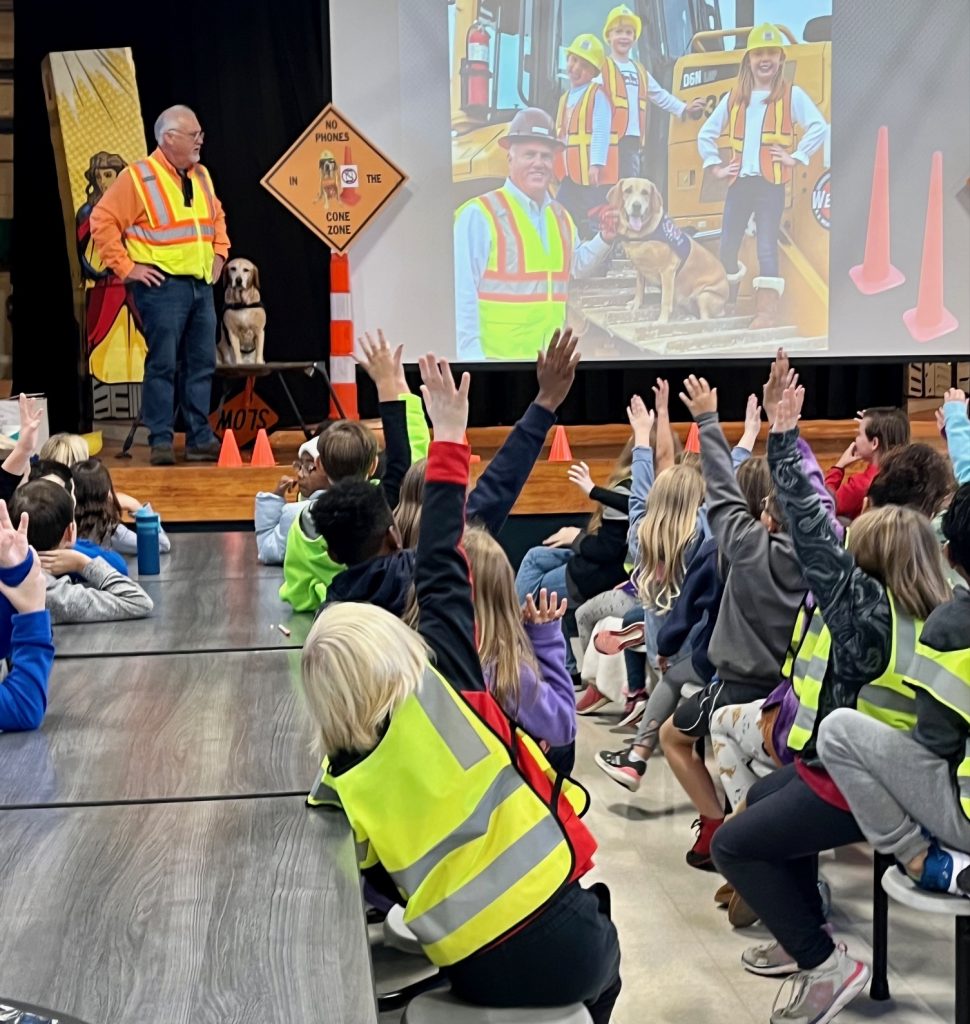 Elementary school age children sitting in a cafeteria raising their hands while Morris King and Millie, a yellow Labrador Retriever, are in the top left corner giving a presentation on work zone safety