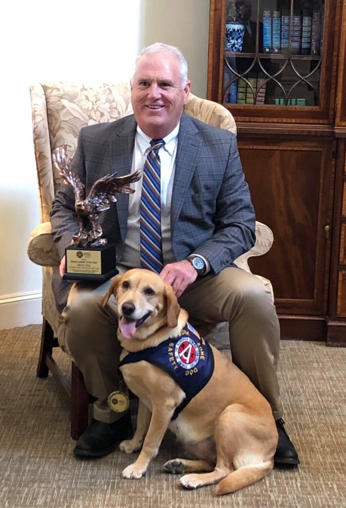 Millie, a yellow Labrador Retriever, poses with her owner Morris King who is sitting down and holding a trophy with a flying eagle on it