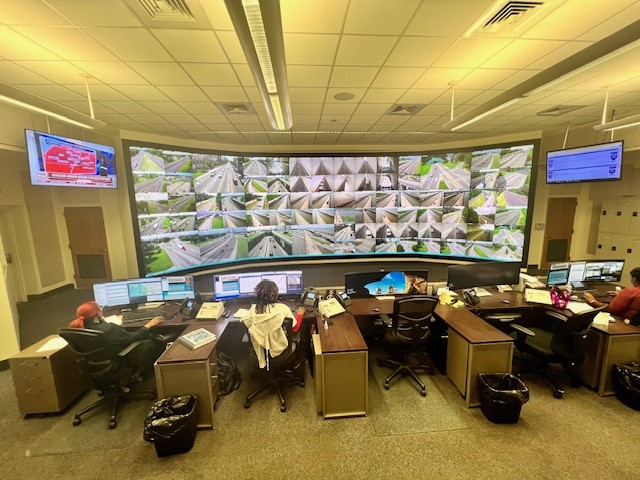 People sitting at desks looking at a giant tv monitor displaying traffic cameras across a region