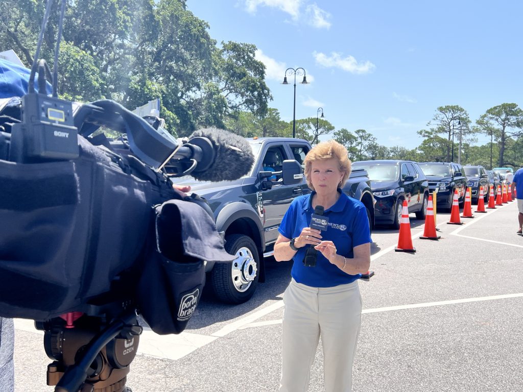 Female reporter speaks into a microphone in front of a line of cars