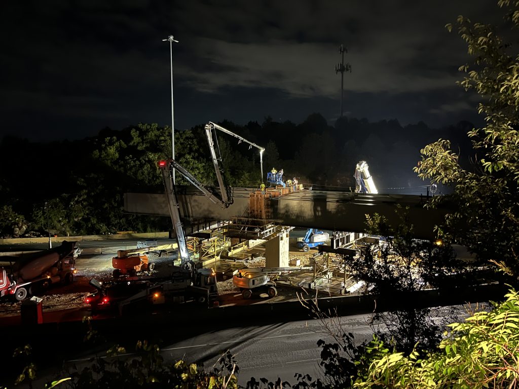 The work area is well-lit at night as workers secure girders spanning the I-565 roadways.