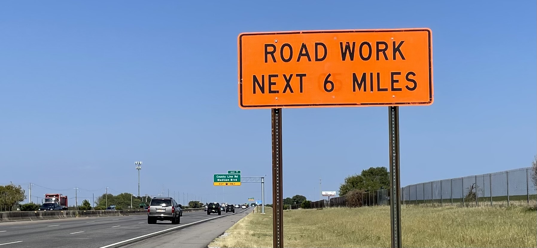 An orange construction sign reading "ROAD WORK NEXT 6 MILES" next to a highway with vehicles traveling on it.