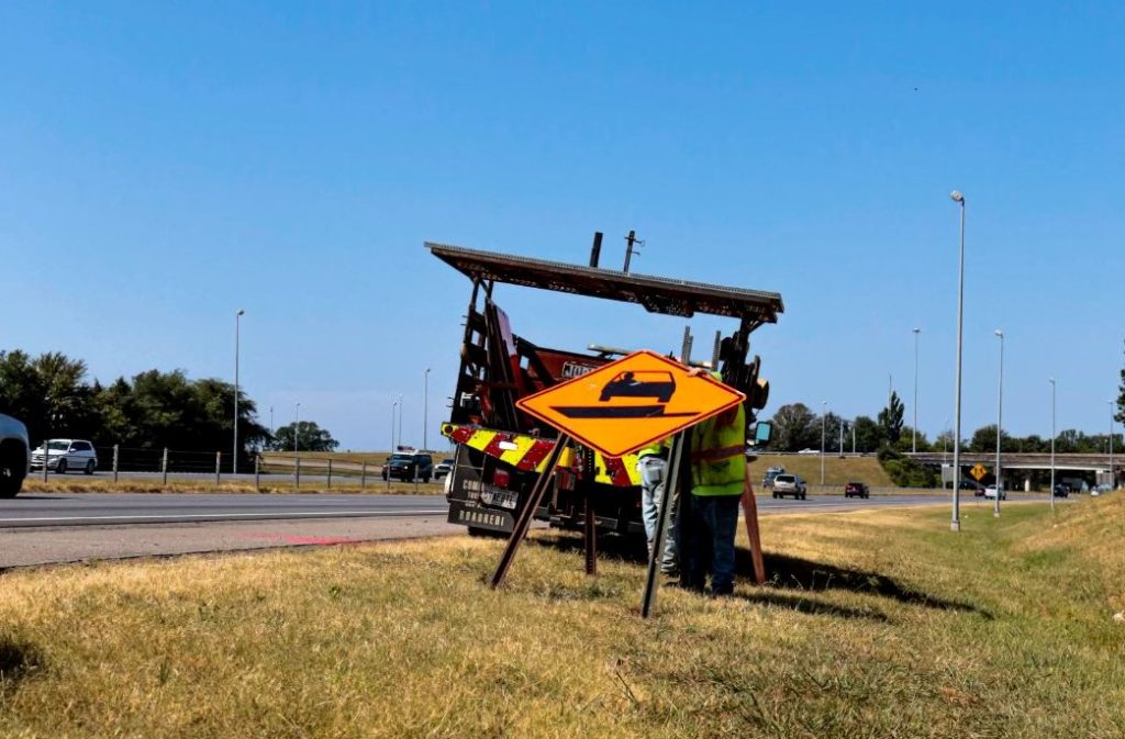 Workers fasten a large orange diamond-shaped sign to posts and prepare to raise it into position.