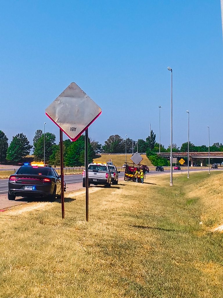 A covered sign next to the highway. A police car, ALDOT trucks, and a subcontractor's sign truck are parked on the shoulder.