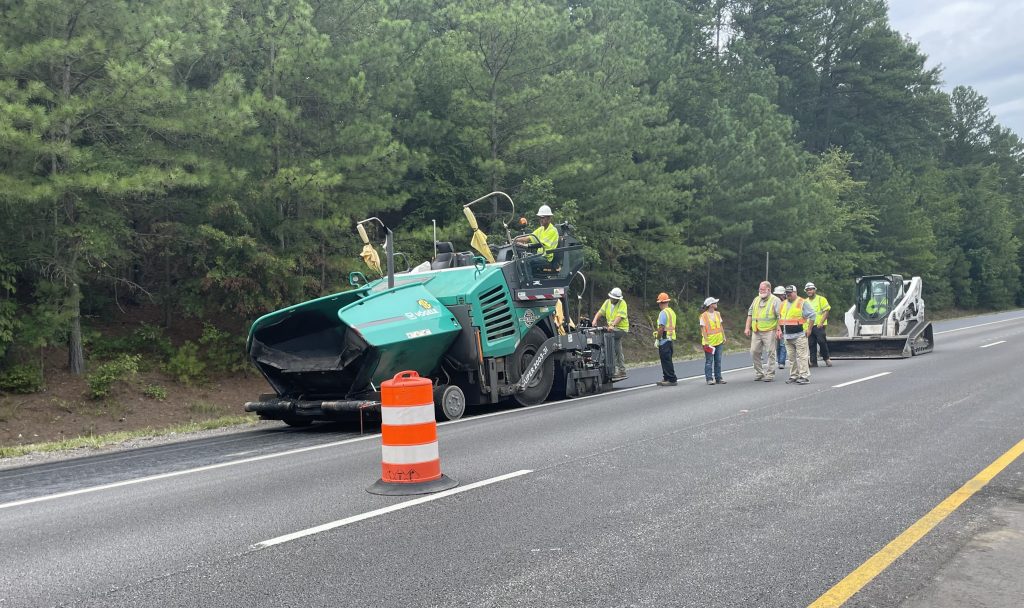 A paving machine moves down the shoulder of the roadway with workers following to smooth any rough areas in the hot asphalt.