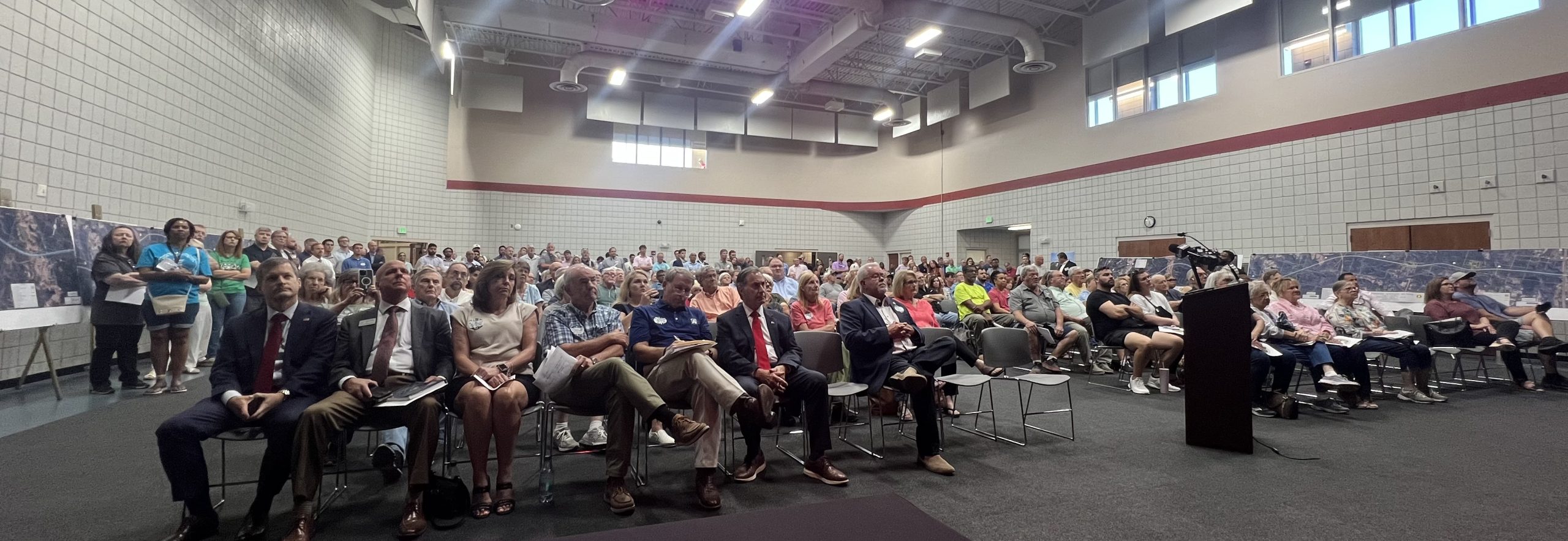 Meeting attendees sitting in chairs facing a stage with a large group of people standing in rows behind the chairs.