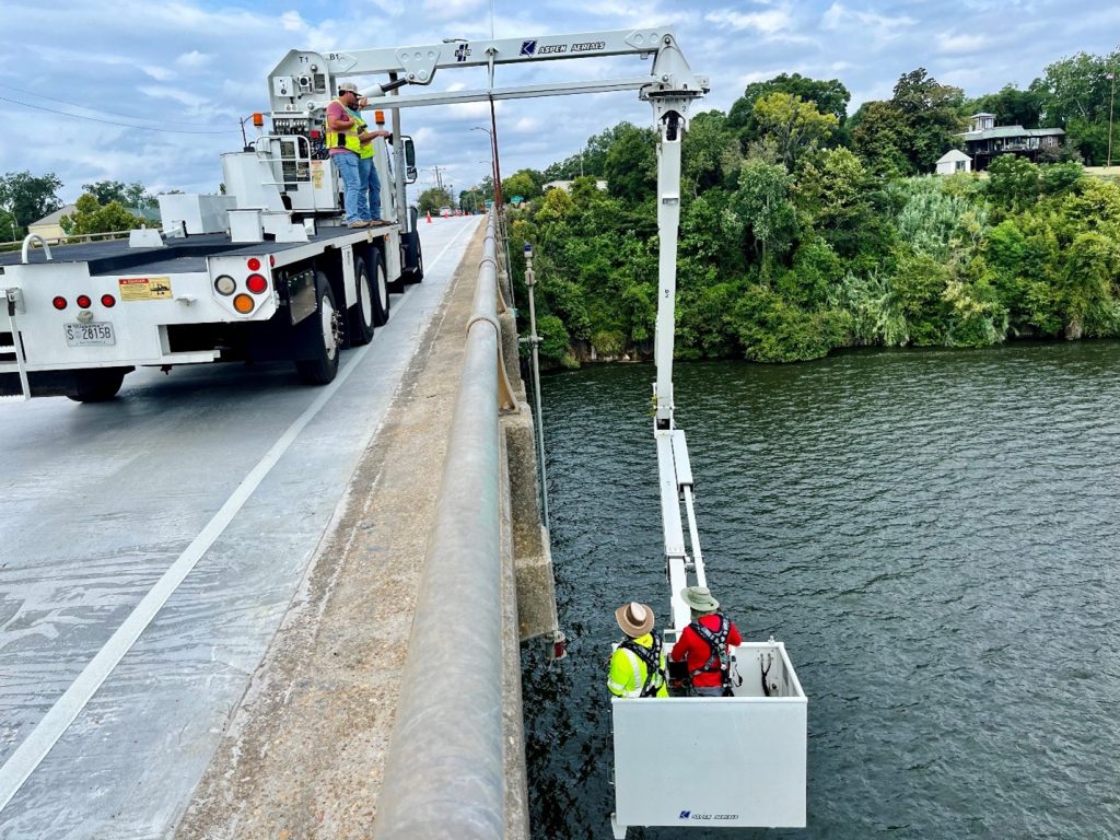 2 people in a bucket inspect under a bridge while 2 people stand on the side on the bridge on the truck and oversee