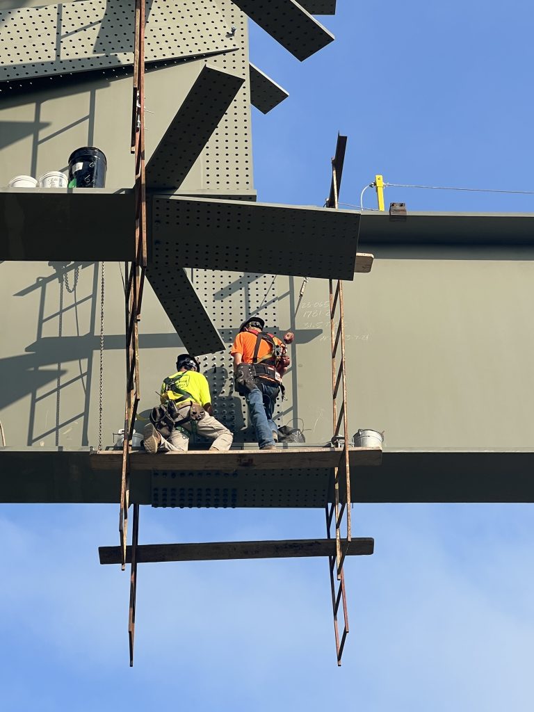 Workers in safety gear work from scaffolding hanging from a bridge girder.