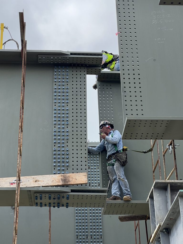 Workers in safety gear reach out to align a suspended beam.