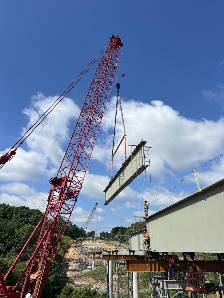 A crane lifts a large steel beam into place next to another beam that has already been installed.