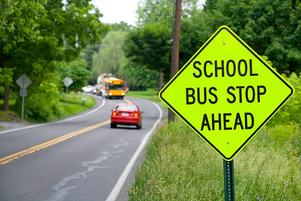 School bus stop sign in new color with bus, car and person. Bus has it's warning lights on. Foreground in focus, car stopping.