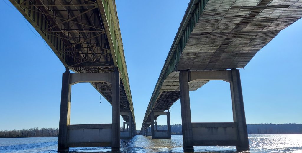 Looking south at the underside of two large bridges over a waterway. The bridge to the right has metal scaffolding suspended under it.