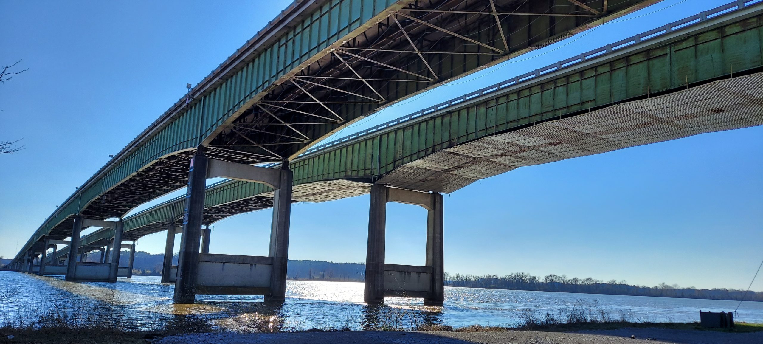 Looking south at the underside of two large bridges over a waterway. The bridge to the right has metal scaffolding suspended under it.