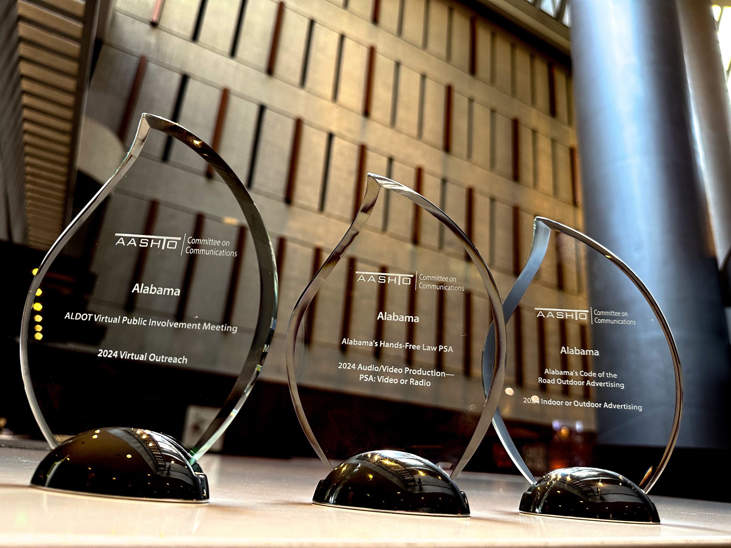 Three glass awards on a table with black bases. Each award has a teardrop shape and is inscribed with text about the Alabama Department of Transportation (ALDOT) and the AASHTO Committee on Communications. The awards recognize ALDOT Virtual Public Involvement Meeting, Alabama's Hands-Free Law PSA, and Alabama's Code of the Road Outdoor Advertising.