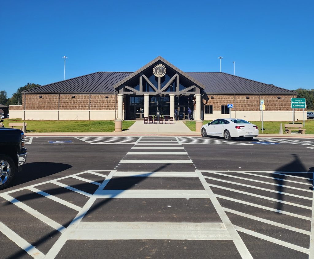 A brick building with a brown metal roof and the Great Seal of Alabama affixed above the entryway, viewed from across a parking lot with a crosswalk leading to the sidewalk.