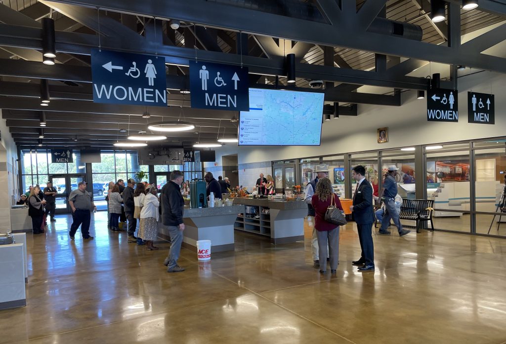 People enjoy refreshments in a large building lobby. A TV screen showing travel conditions in the state hangs overhead.