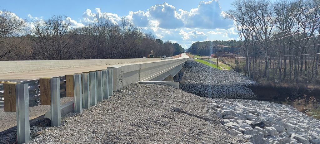 A new concrete bridge with shiny guardrail approaching it, over a creek with riprap stone lining the slopes.