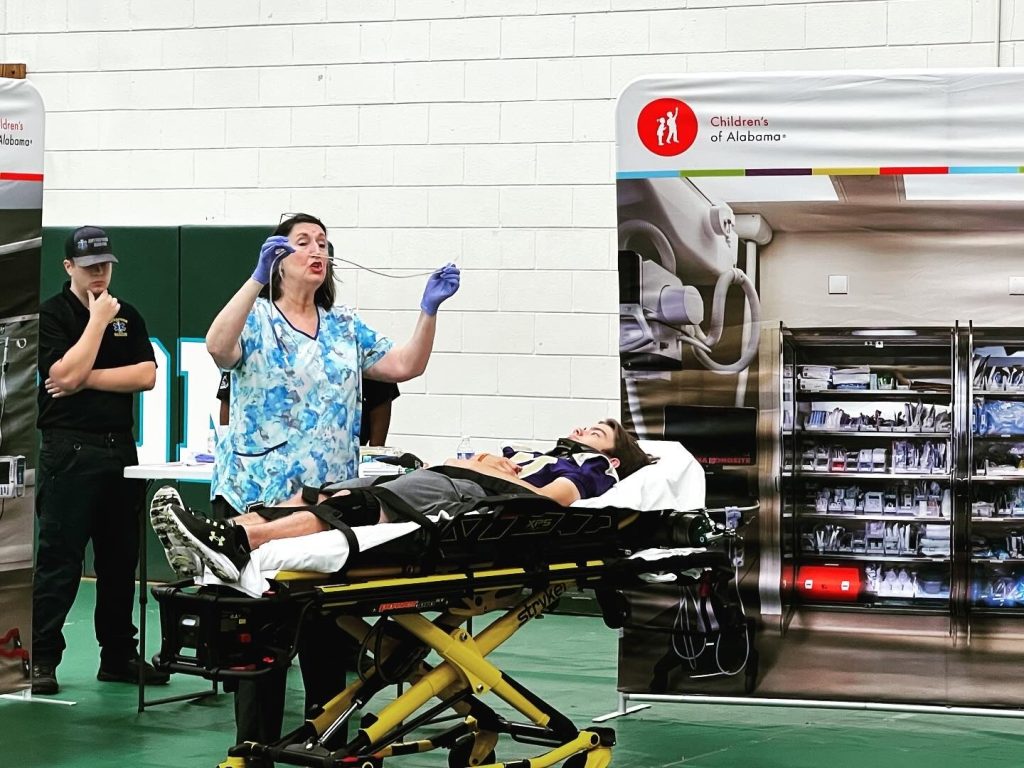 Nurse in a gym showing crowd out of frame a tube. Student lying on gurney in neck brace.