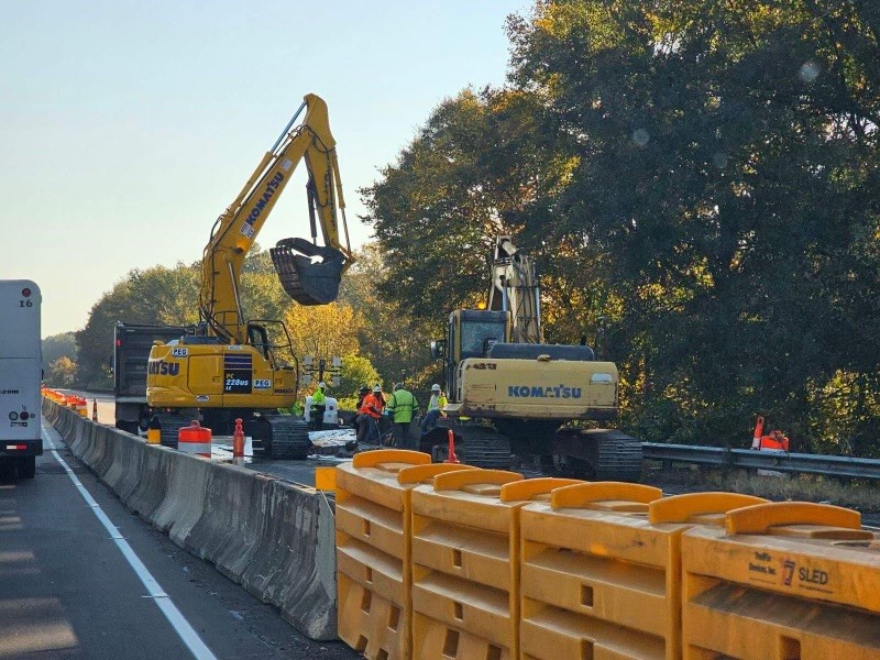 A yellow construction excavator is digging into the road surface while workers and equipment are visible on the side of the highway. Traffic cones and barriers are in place to protect the workers.