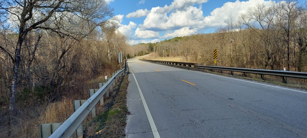 A road and a bridge, with trees and trees and sky in background.