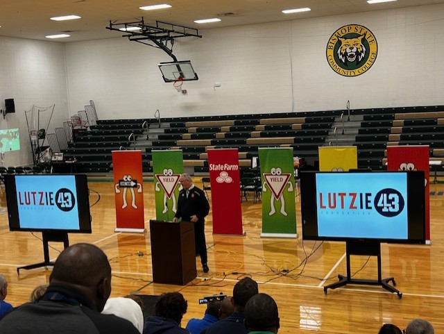 High school gym, one way, yeild and stop signs in background. Man standing at podium giving speech to crowd.