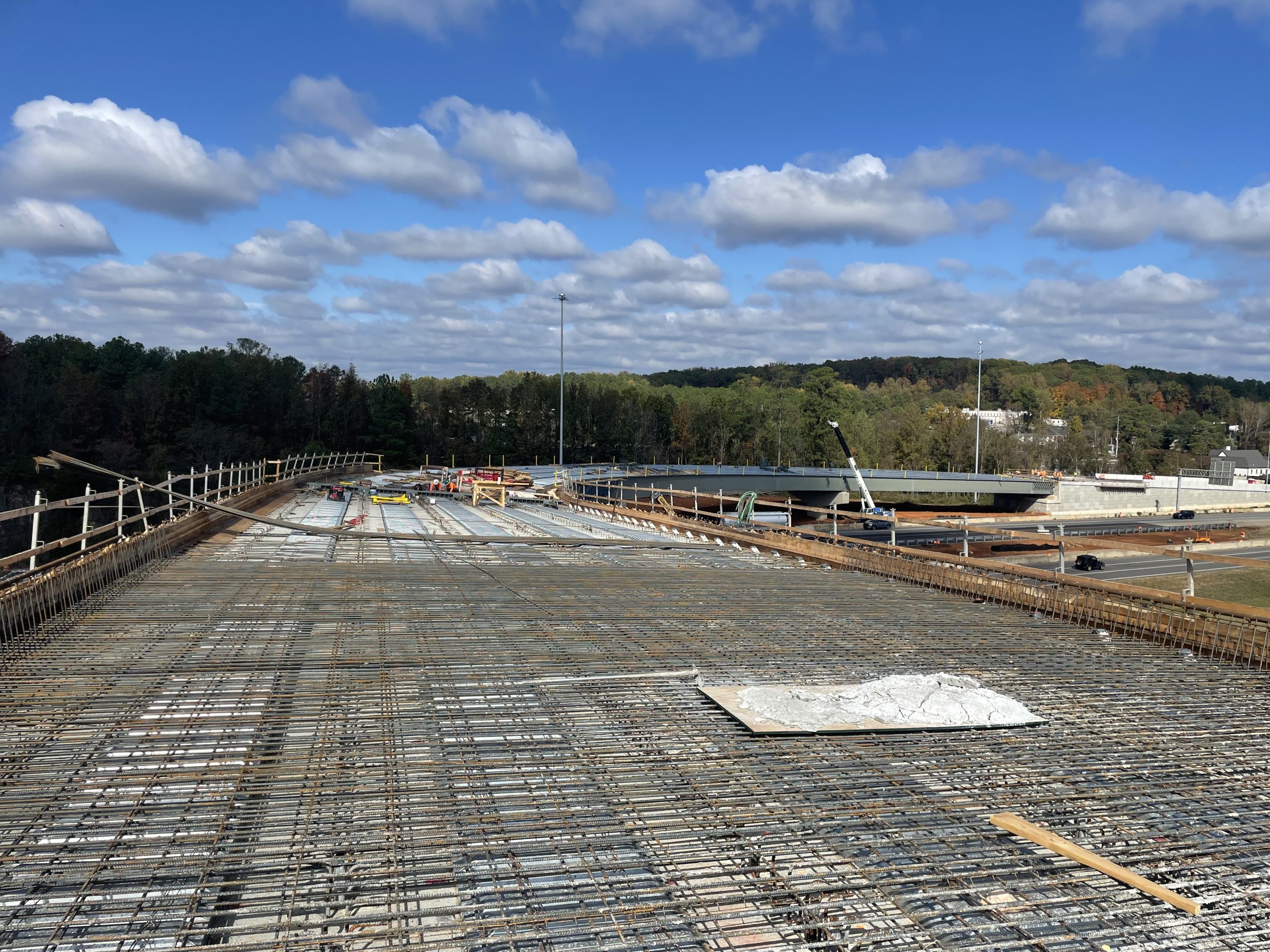 The image shows a large construction site with a bridge under construction. The bridge deck is visible, covered in a grid of rebar. Construction equipment and workers are visible in the background. The sky is blue with some white clouds.