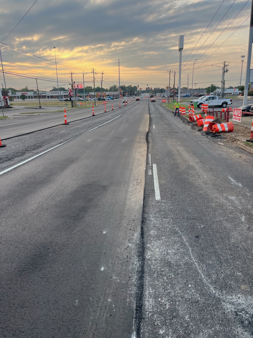 A stretch of road under construction. The asphalt has been freshly laid on the right side of the road, while the left side remains in its original condition. Orange traffic cones and barrels line the sides of the road, indicating ongoing work. The sky is overcast with a hint of a sunset in the distance.