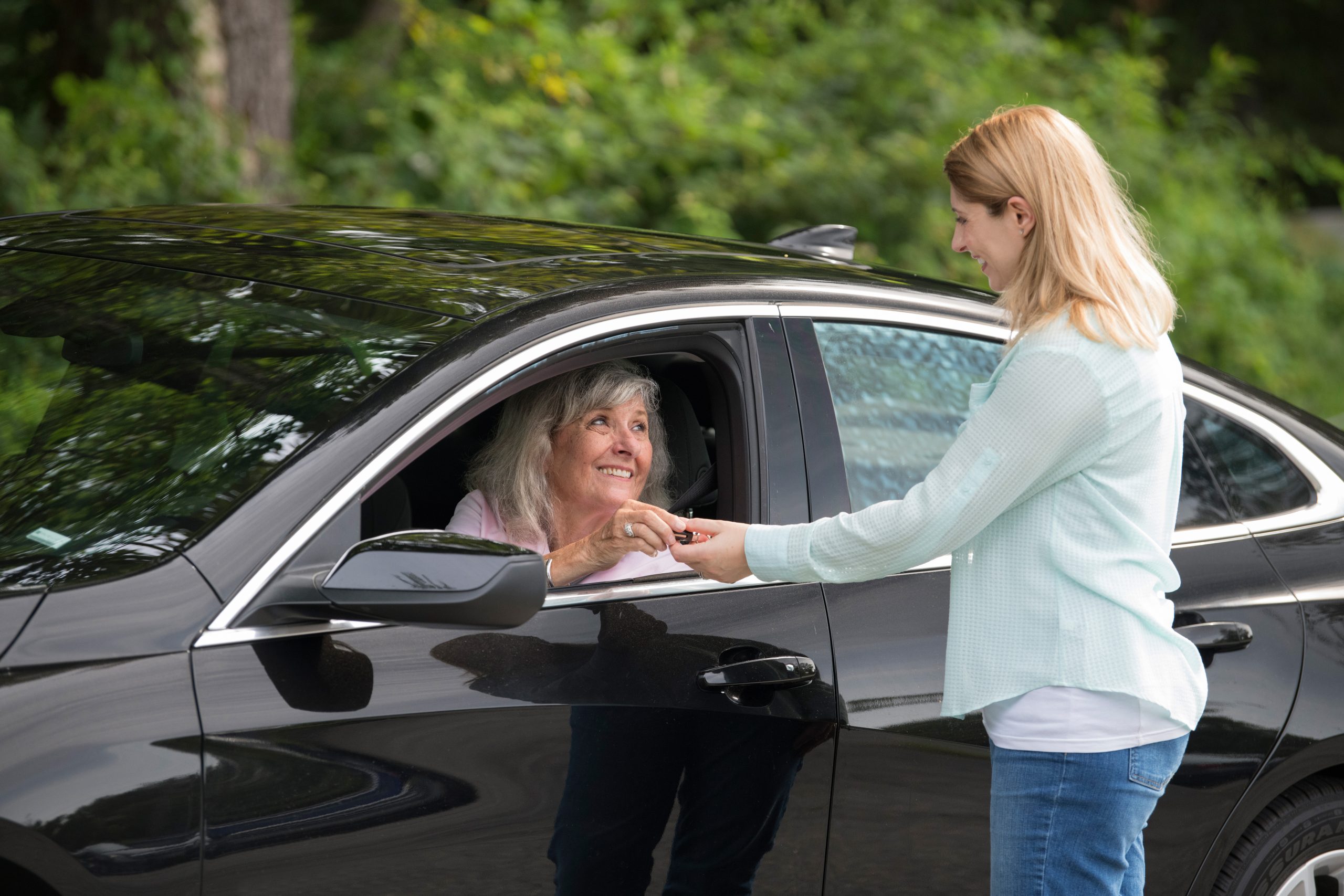 A black car with an older driver and blonde hair lady in a blue shirt handing the older drive a key to the black car.