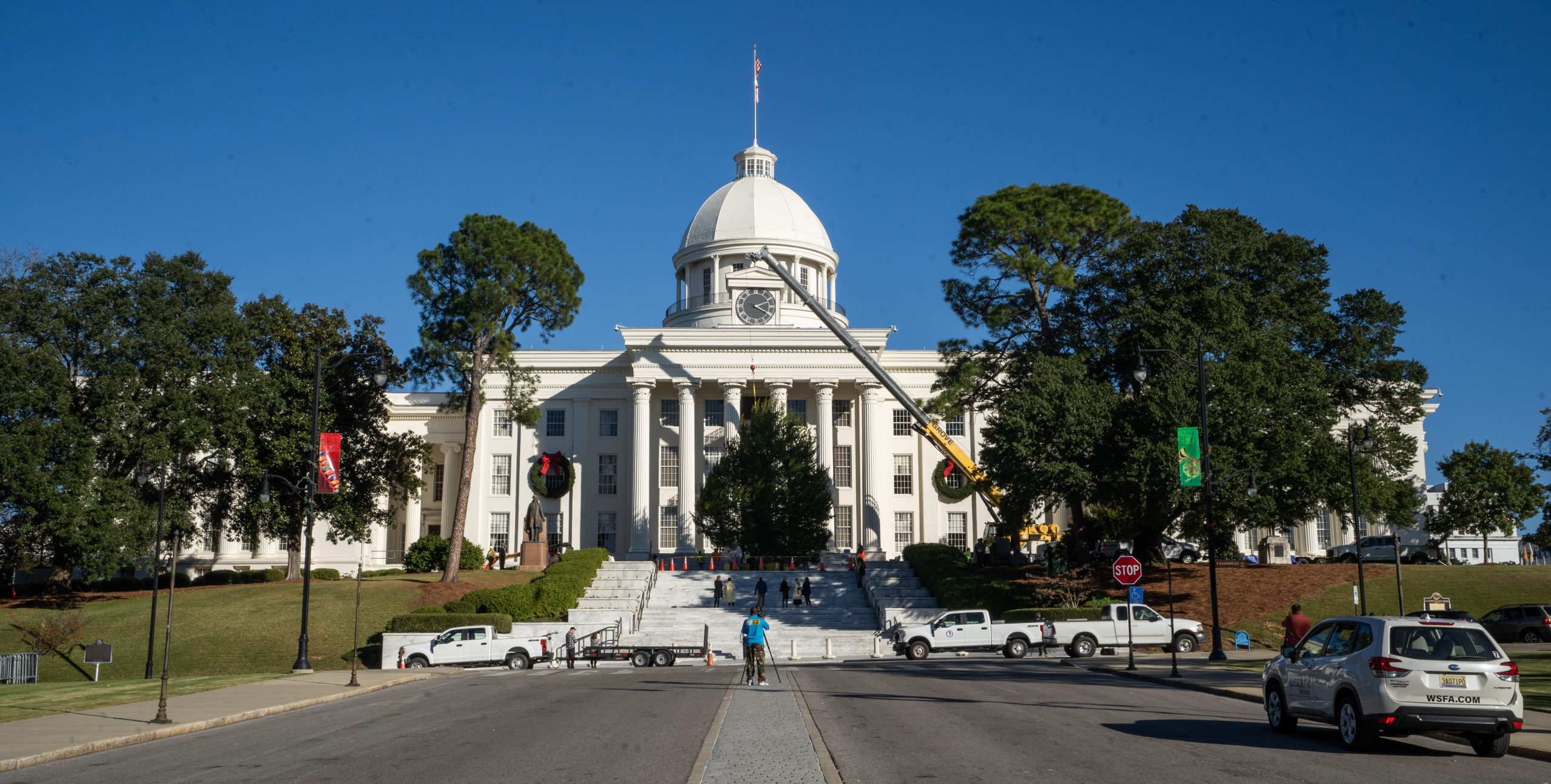 The Alabama State Capitol building in Montgomery, Alabama, with large Christmas wreaths hanging on the windows and a crane lifting a christmas tree to its place on the steps