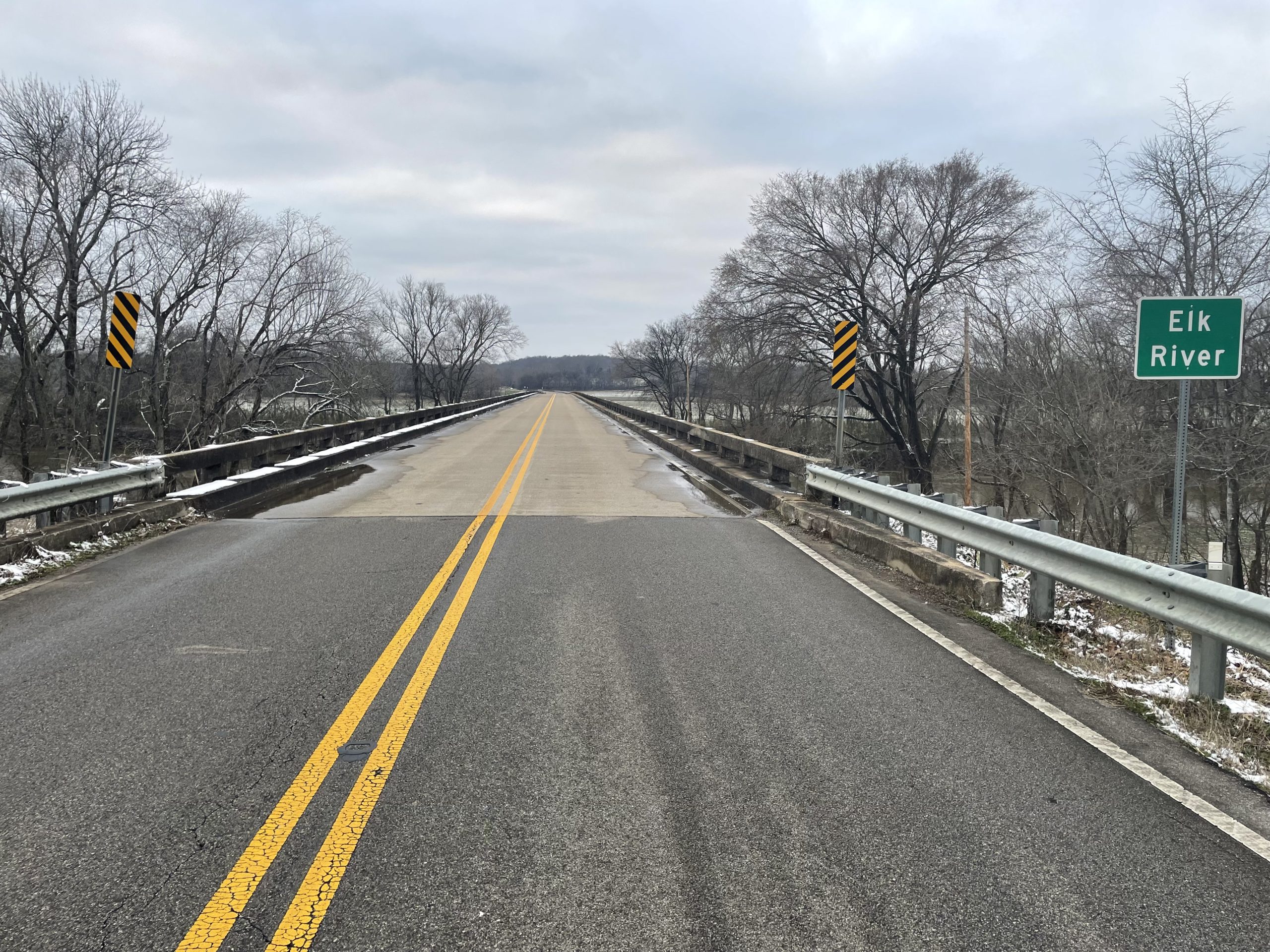 A roadway entering a bridge with a green sign reading "Elk River" in white lettering