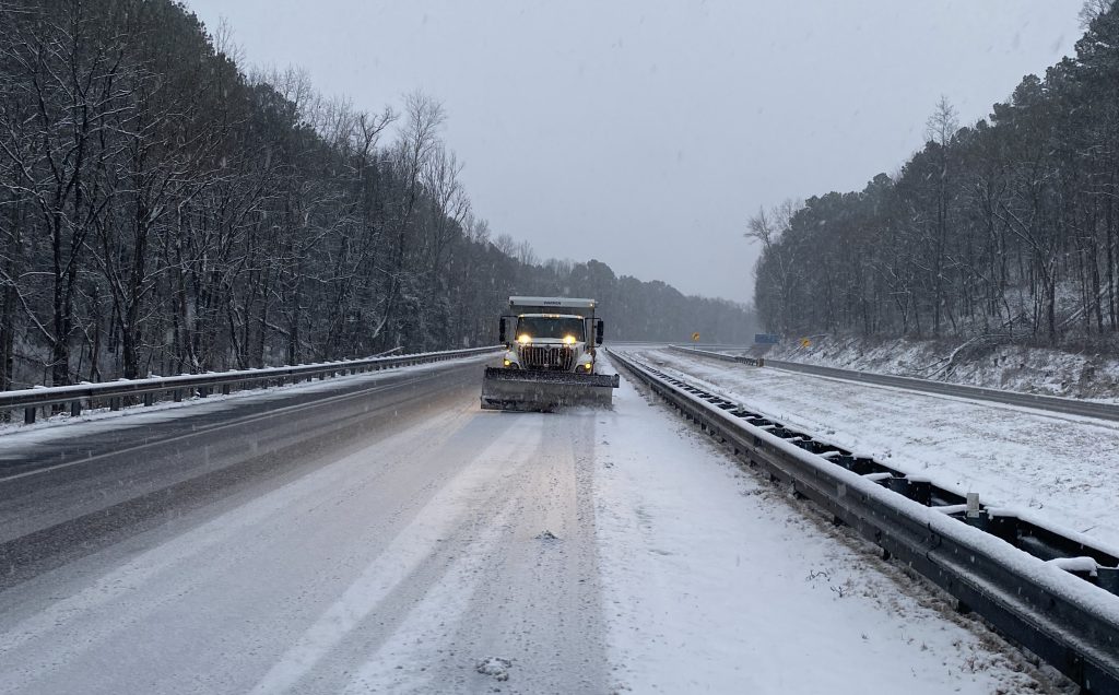 An ALDOT truck with plow clearing snow-covered interstate travel lanes between guardrails.