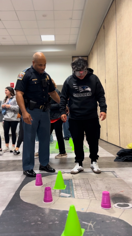 State trooper helping student stand up straight with drunk goggles on
