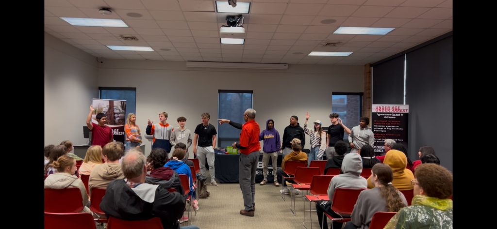 Man in red shirt pointing to students standing in front of a room.
