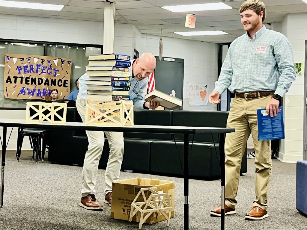 Two men are testing the strength of small wooden model of a bridge by stacking books on them in a classroom.