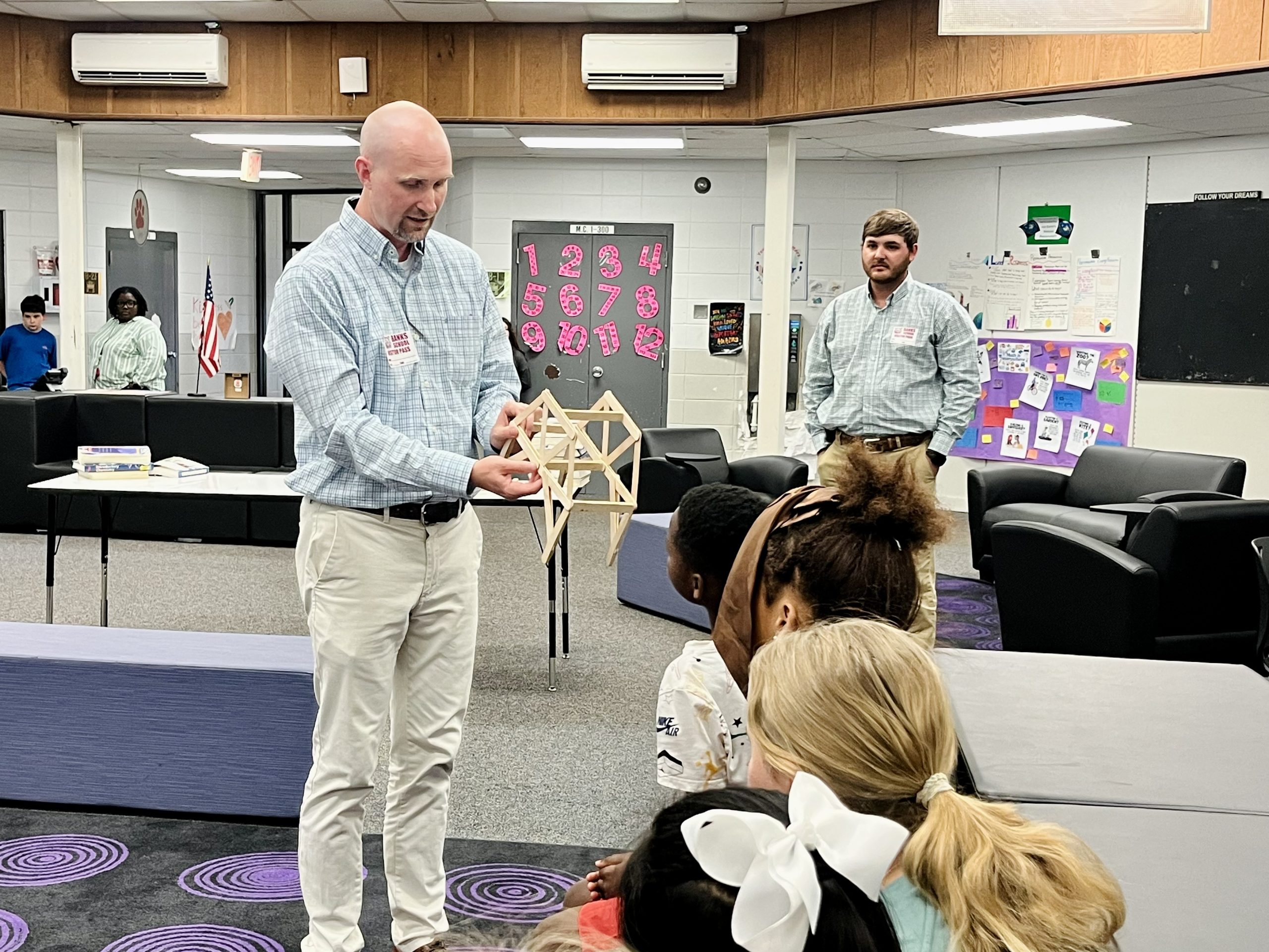 A man in a light blue patterned shirt and khaki pants holds up a small wooden model bridge for a group of seated children in a classroom. Another man stands in the background.