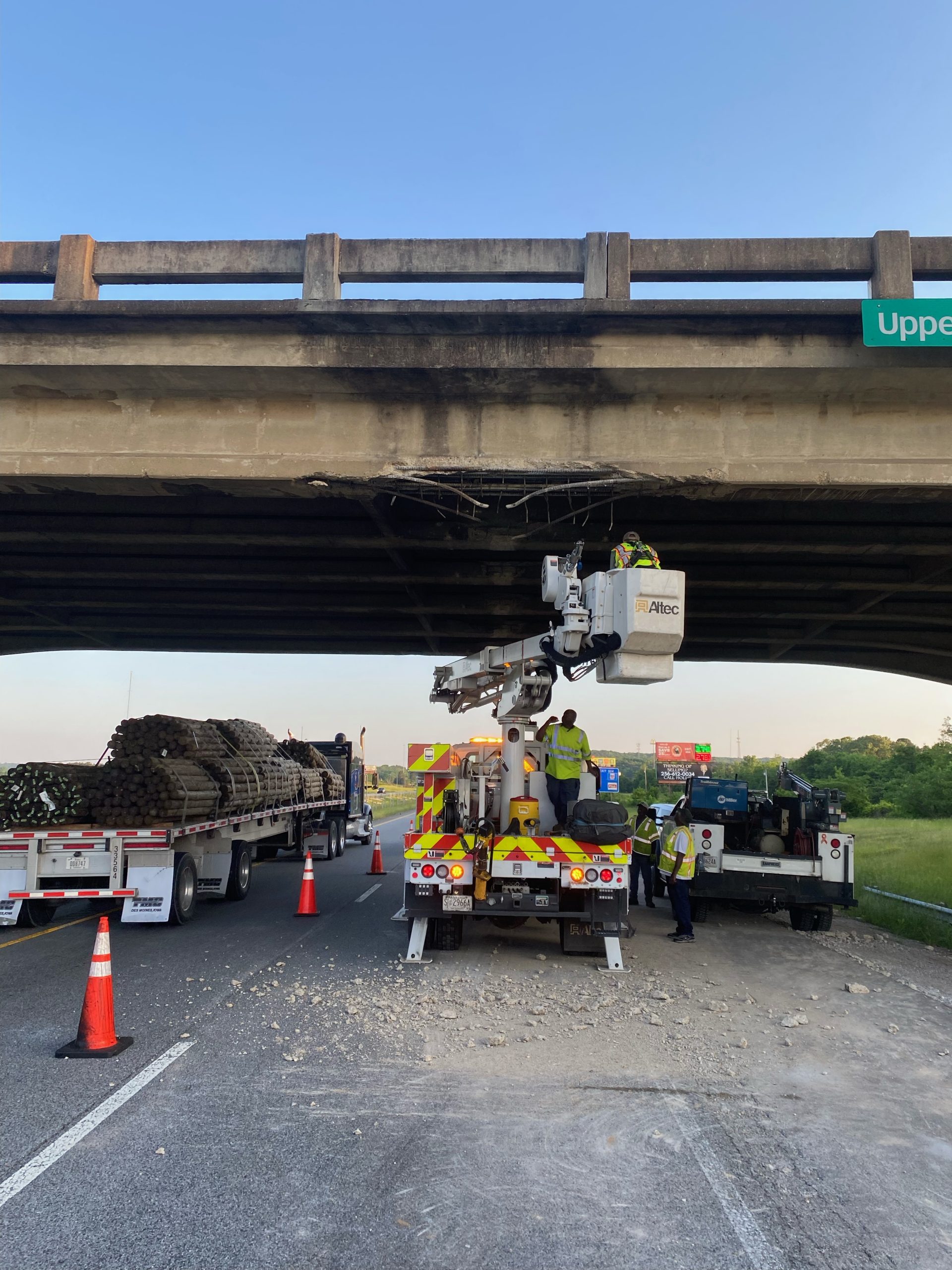 A bridge with a bucket truck underneath. A worker in a yellow vest and harness is in the bucket. Loose concrete debris litters the roadway.