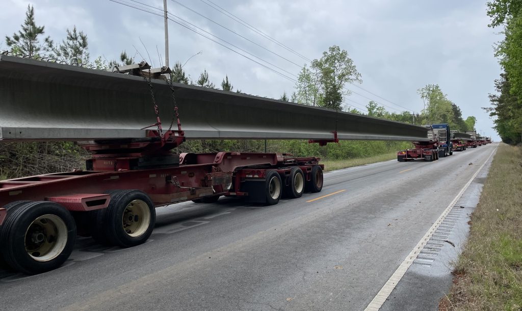 Several trucks, each hauling a massive concrete beam, are lined up on the road.