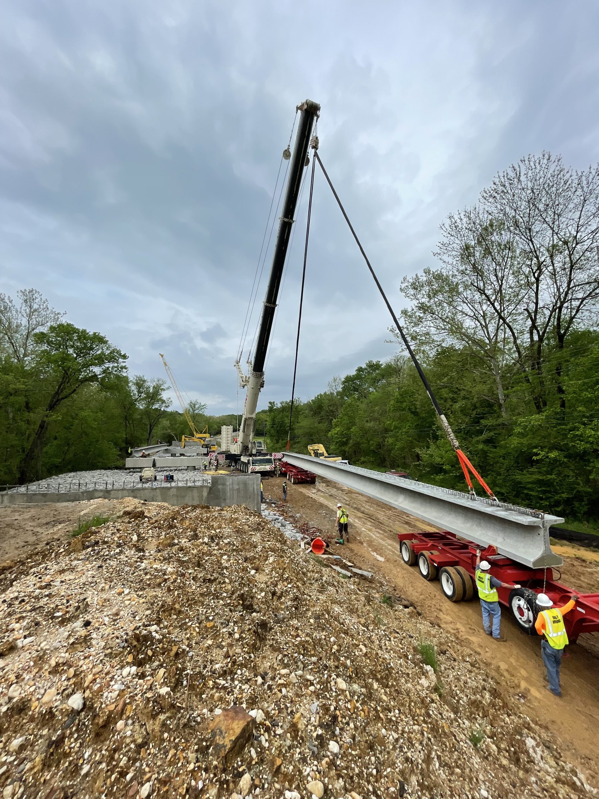 A crane with cables hooked to a long concrete beam. Workers in yellow vests and hard hats stand by to assist with guiding the girder as it is moved.