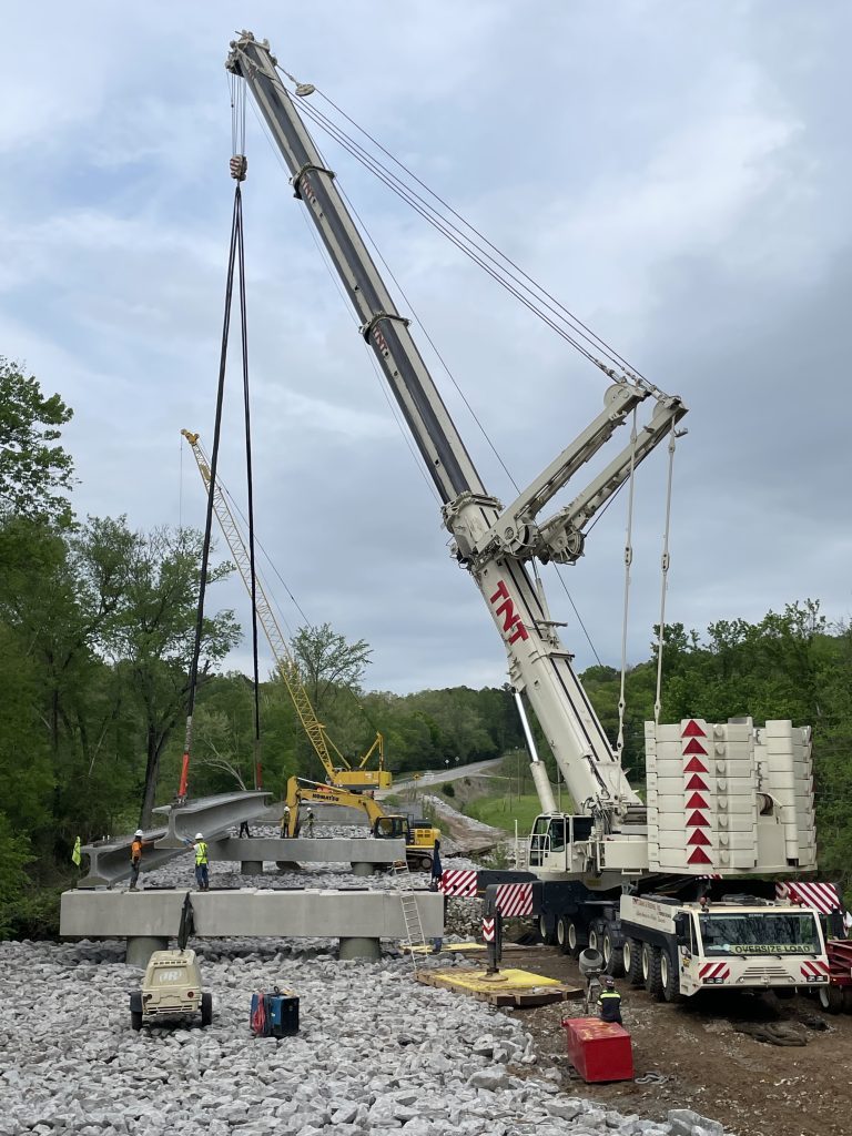 A bridge girder, suspended from a crane, being lowered onto the bridge caps. Workers in yellow vests and hard hats help direct it's positioning.