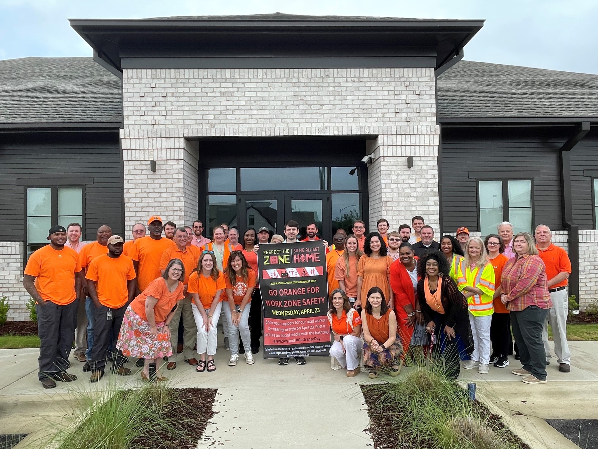 ALDOT West Central Region employees, wearing orange, pose in front of a building holding a work zone awareness week picture