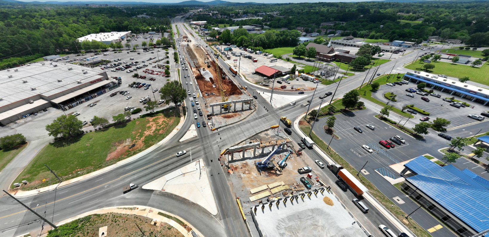 An aerial view of an intersection of two roads. The major route has a wide median area that is under construction for a new highway mianline. Bridge columns are being built near the intersection.