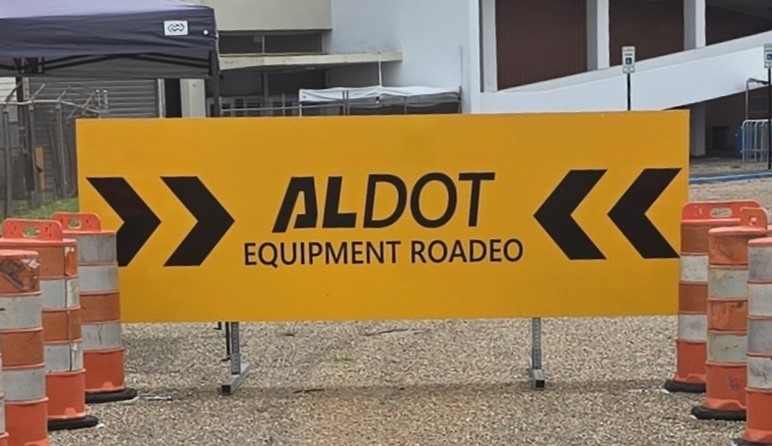 Yellow sign that reads ALDOT Equipment ROADEO with orange and white construction barrels on each side.