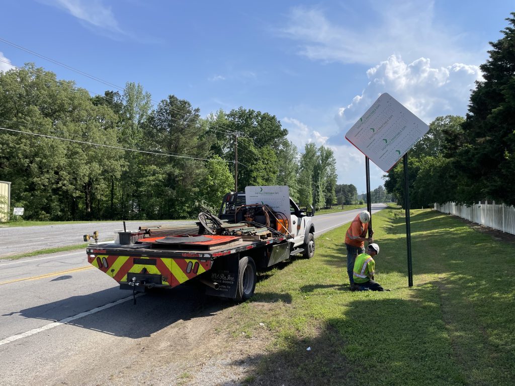 Truck with signs on it and two workers in hardhats installing a covered sign on the highway shoulder