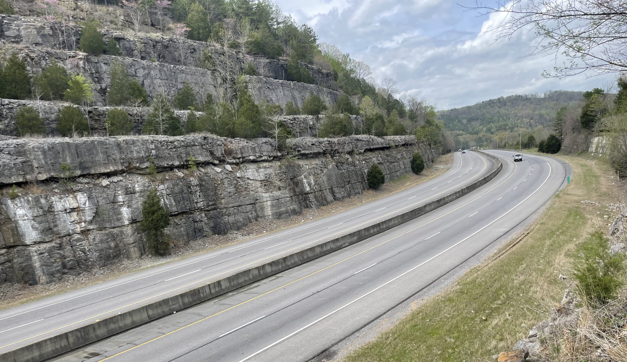 A divided four-lane highway with a median barrier passing through a rock cut.
