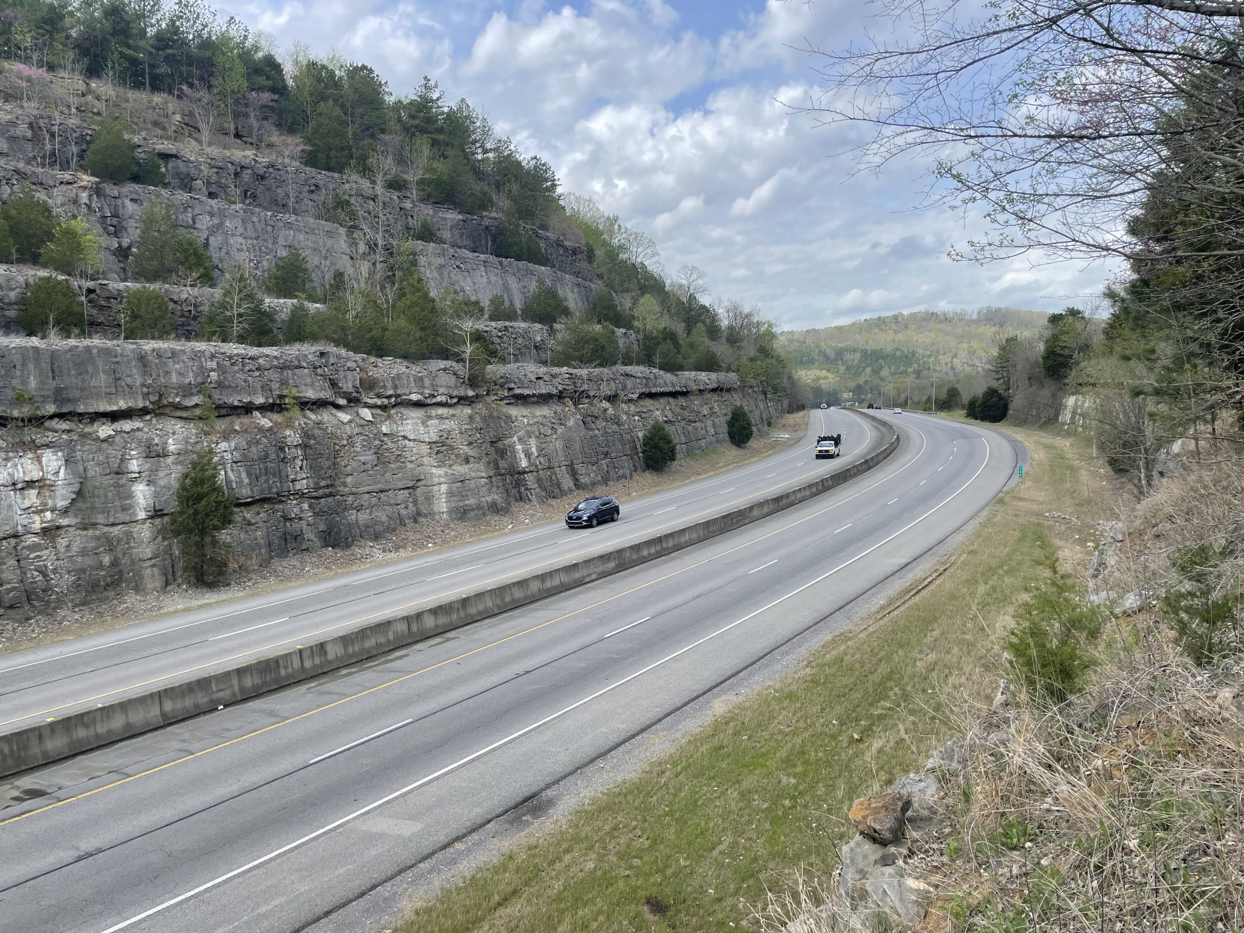 A divided four-lane highway with a median barrier passing through a rock cut.