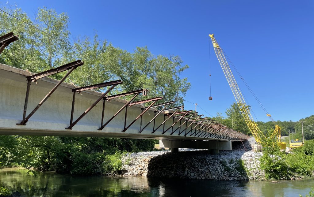 A bridge in an intermediate stage of construction, with girders visible but the deck unfinished and no rail. Water flows beneath. A crane looms in the blue sky on the other side of the creek.