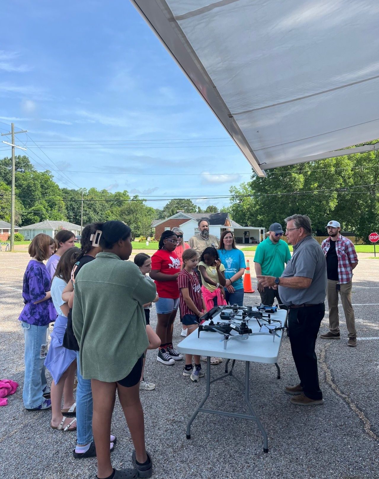 An ALDOT UAS team member speaks to a group of students gathered around a table displaying drones during an outdoor STEM camp. The group stands under a canopy in a parking lot with trees and houses in the background.