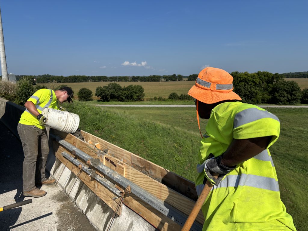 One man in a yellow vest pours a bucket of concrete into a wooden form while another holds a shovel