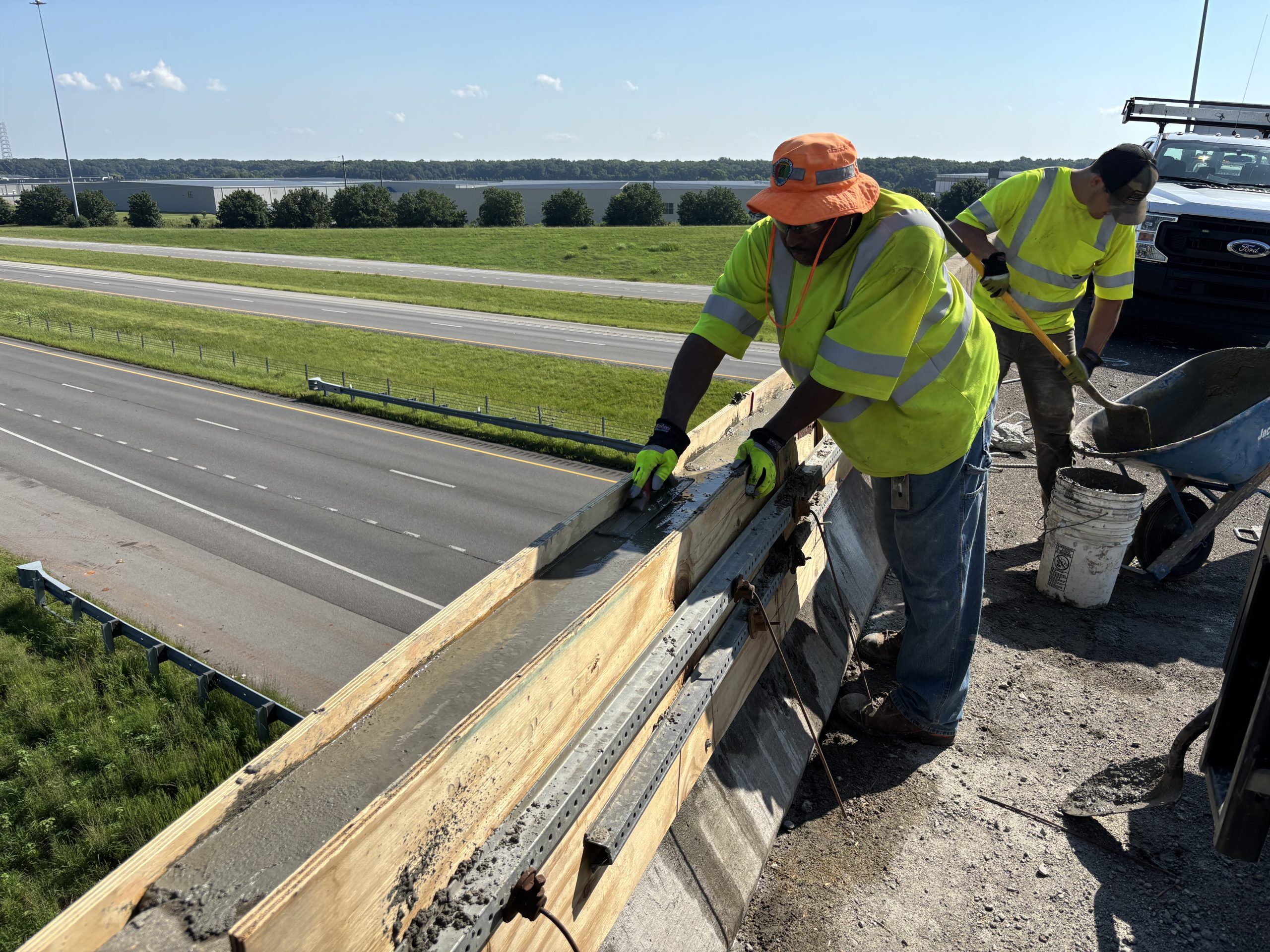An ALDOT worker smooths wet concrete with a trowel while another mixes concrete in a wheelbarrow